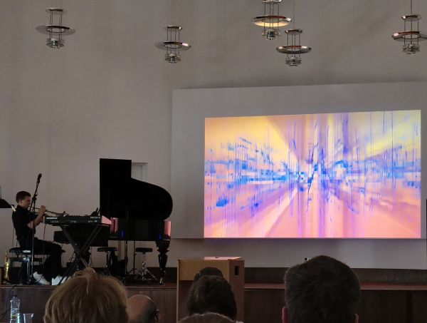 Musiker sitz am Klavier und spielt Trompete, Leinwand zeigt bunte Querlinien